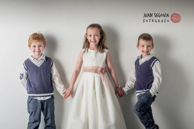 Fotografía de Comunión en Familia - Fotógrafo de Tarragona Fotografía en estudio de tres hermanos sonrientes cogidos de la mano para reportaje de comunión en familia, por Juan Segovia fotógrafo en su estudio de Tarragona